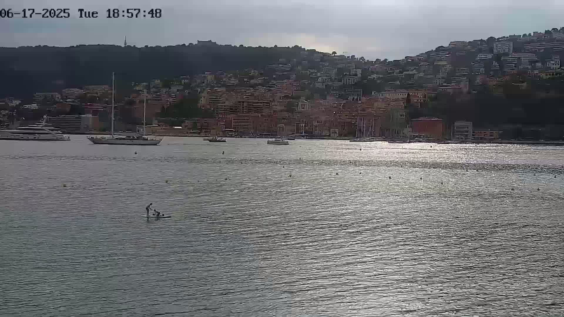 A calm, glistening body of water shows two people on a paddleboard in the foreground, with several boats moored in a harbor and a hillside town in the background under a cloudy sky.