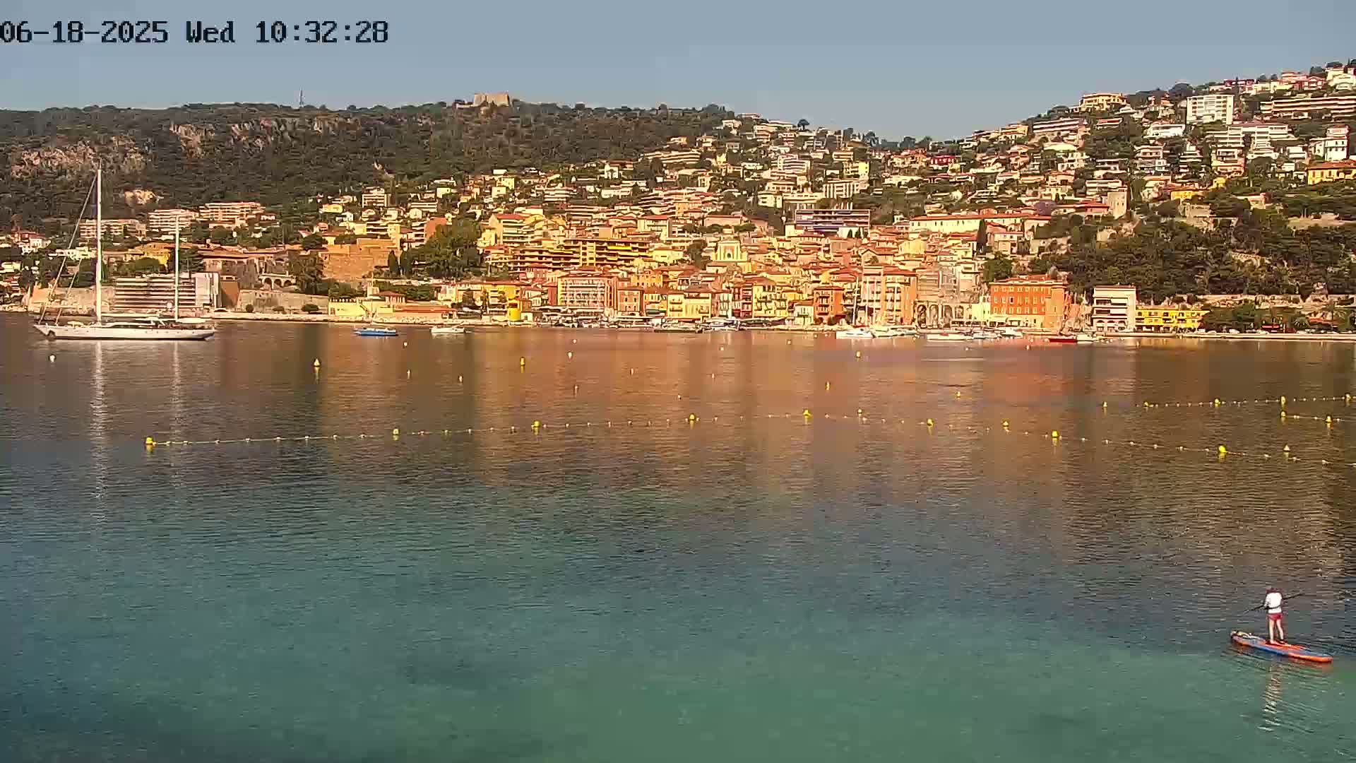 A person paddleboards in calm, clear water in front of a hillside town of colorful buildings on a sunny day.