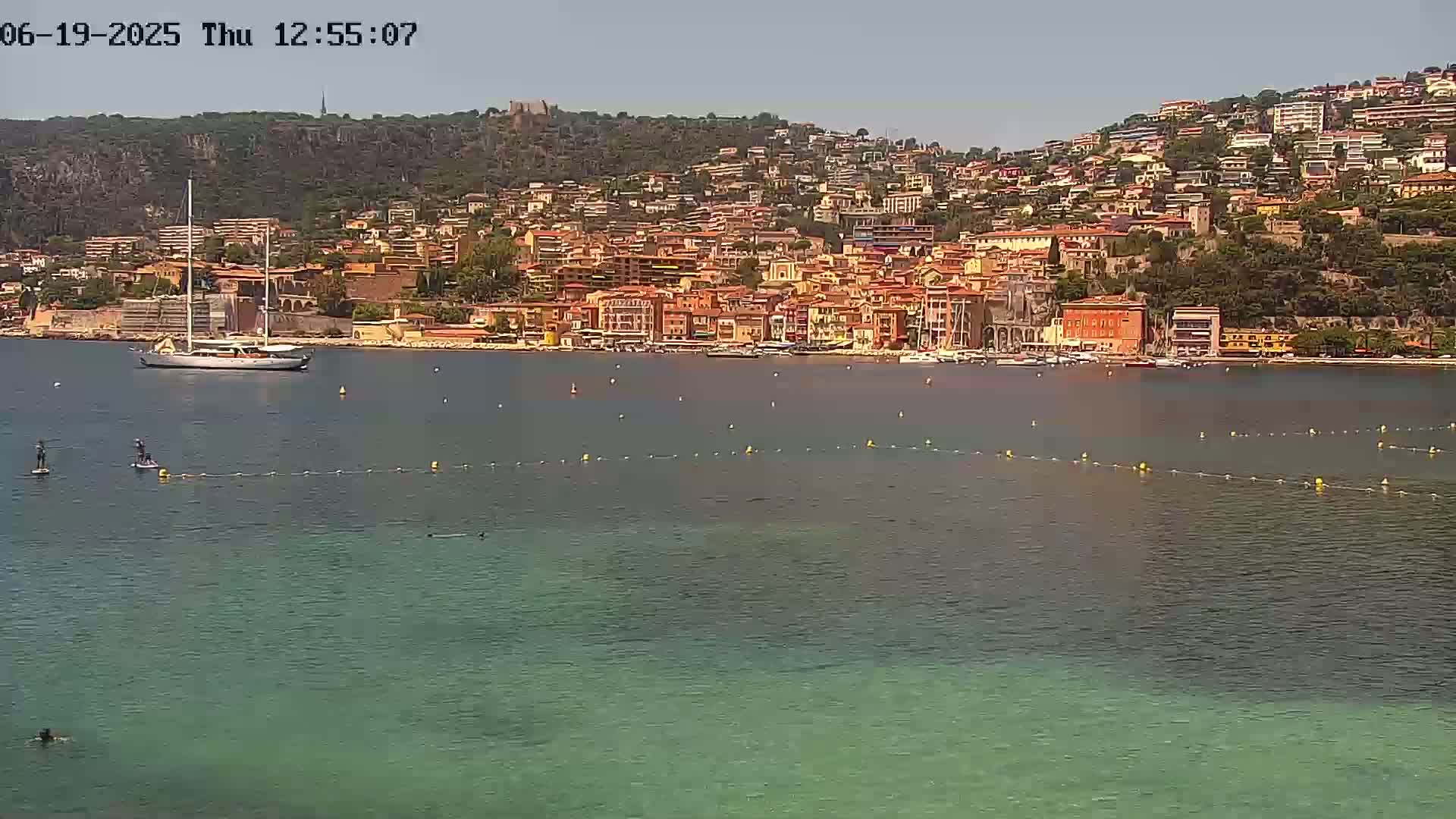 A sailboat is anchored in a calm bay, in front of a hillside town of orange and red buildings under a clear sky.