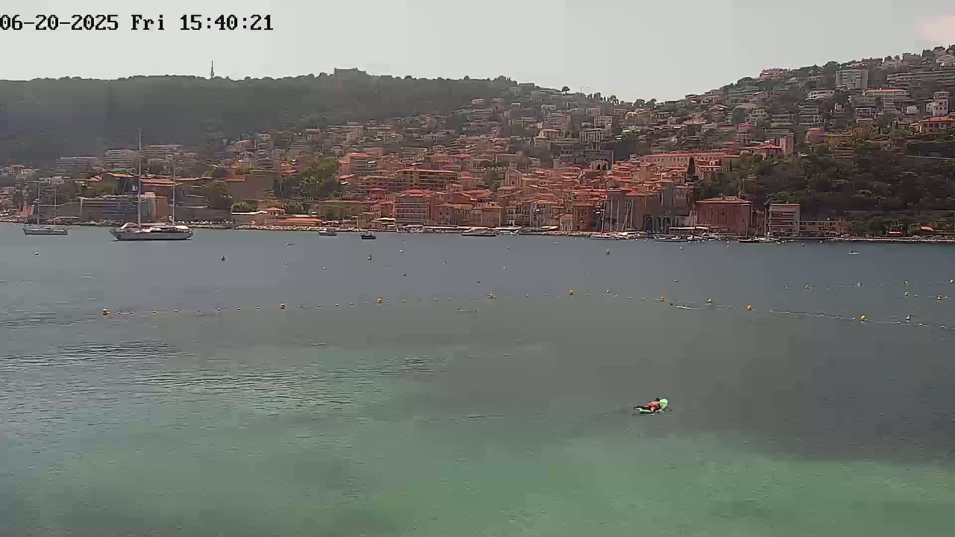 A calm, hazy day reveals a coastal town nestled on a hillside overlooking a tranquil bay with a person paddleboarding in the foreground.