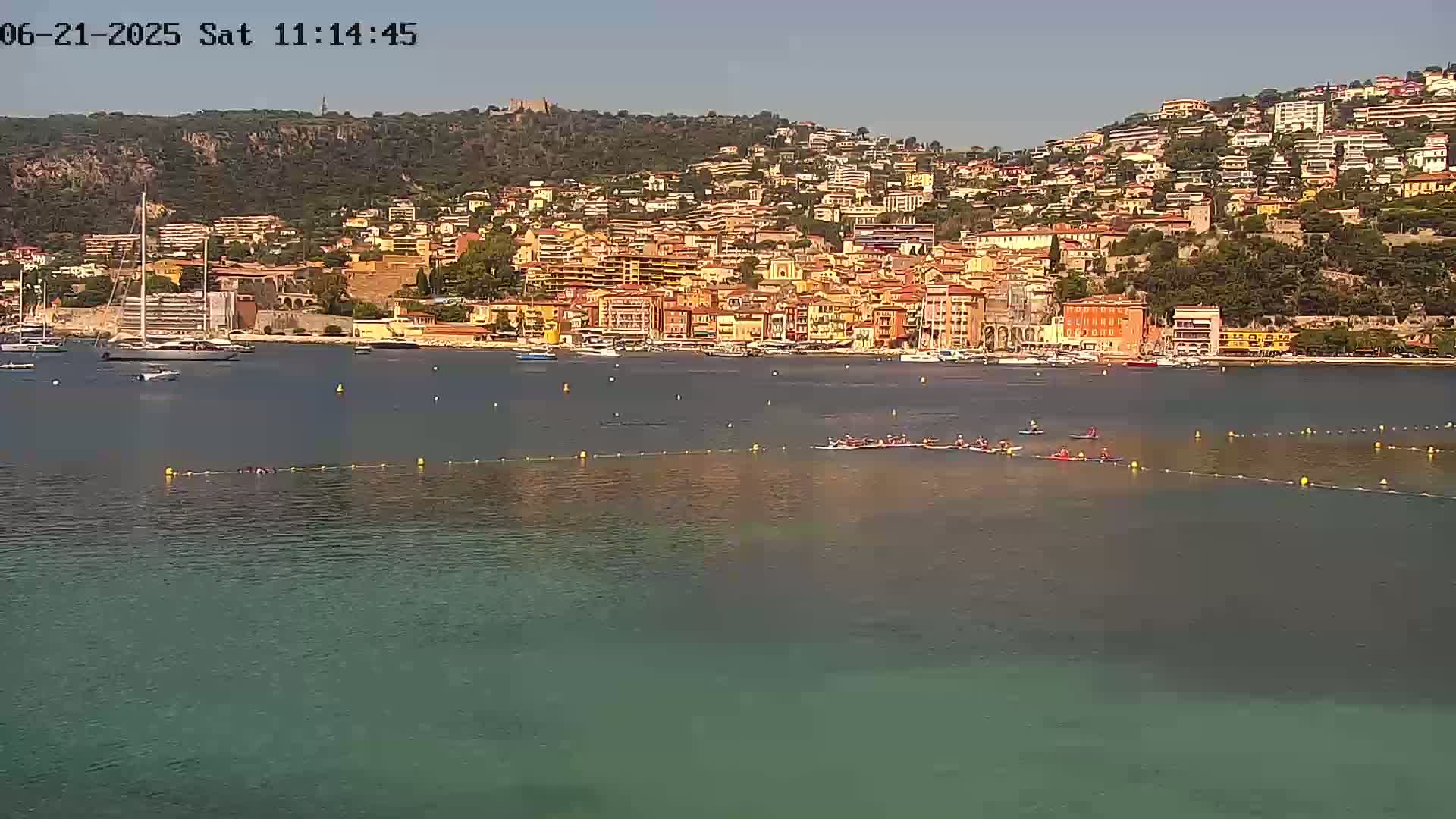 A coastal town with numerous buildings nestled on a hillside overlooking a calm, teal-colored bay where several people are participating in a water sport.