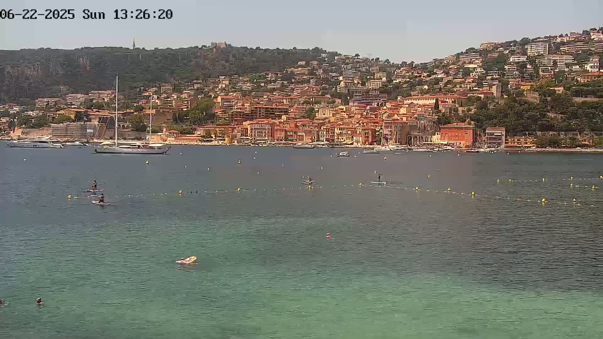 A calm body of water with paddleboarders and a few swimmers in front of a hillside town under a clear sky.