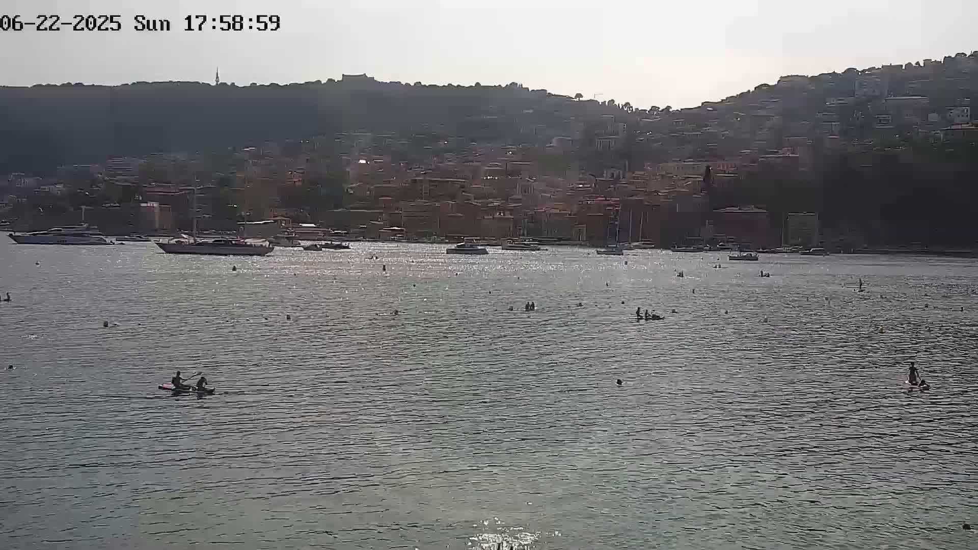 A calm body of water with several people swimming and paddleboarding in front of a hilly town under a bright, sunny sky.