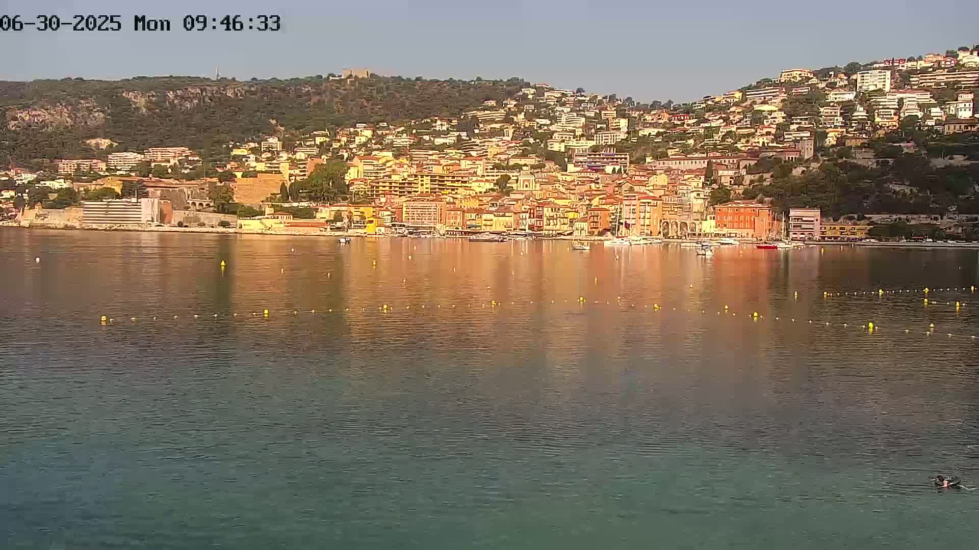 A calm body of water reflects a hillside town of closely packed buildings under a clear sky.