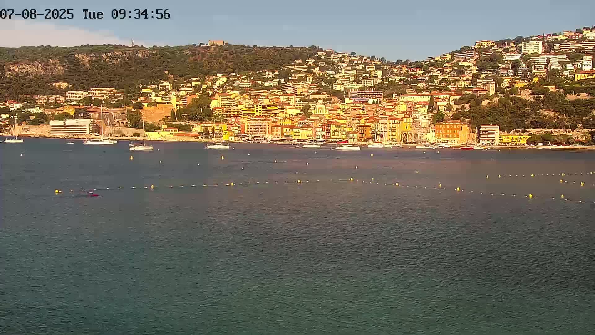 A calm body of water with several sailboats and a hilly coastal town of colorful buildings in the background under a clear sky.
