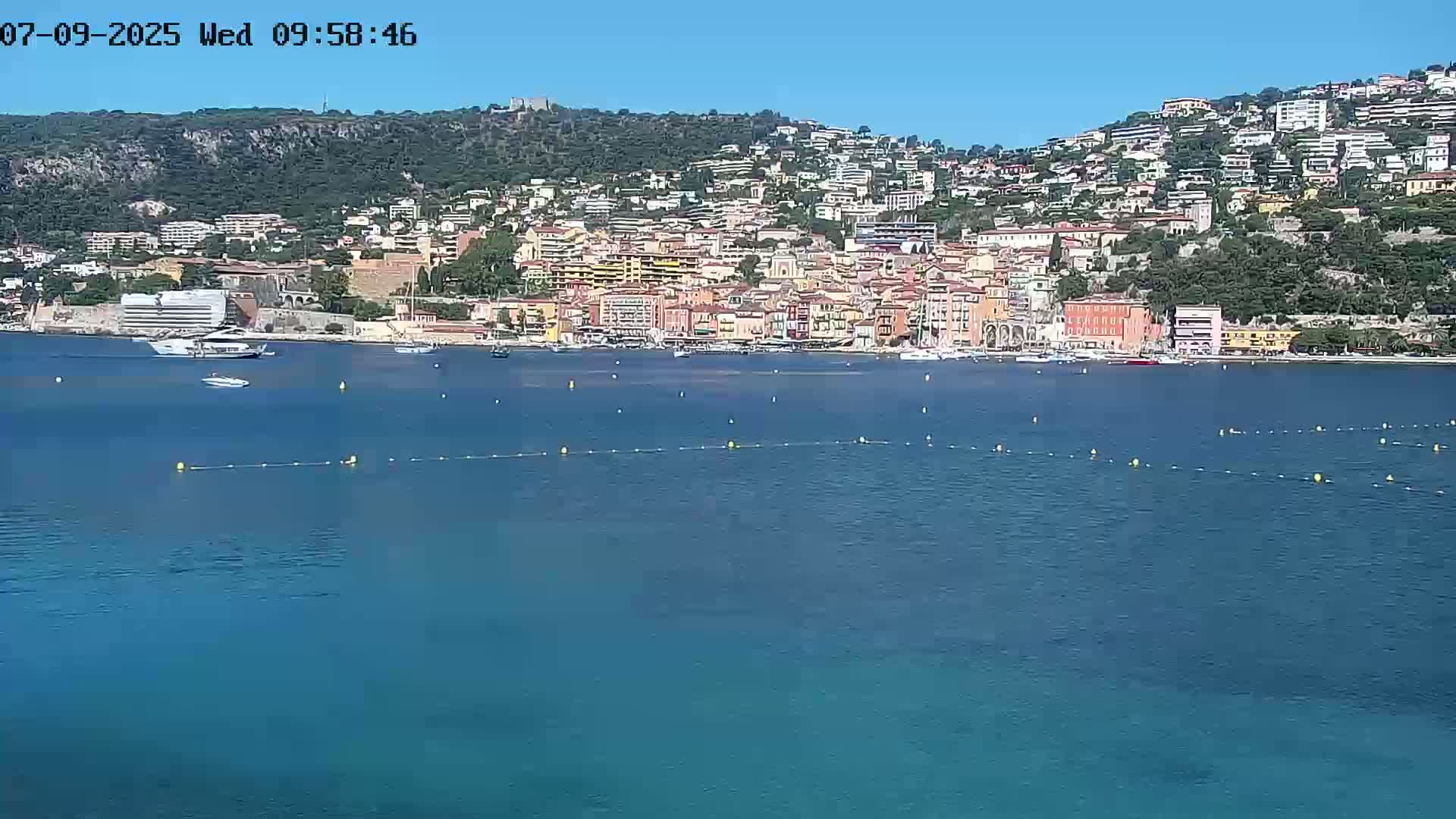 A coastal town with numerous buildings on a hillside sits across a calm, blue body of water under a clear sky.