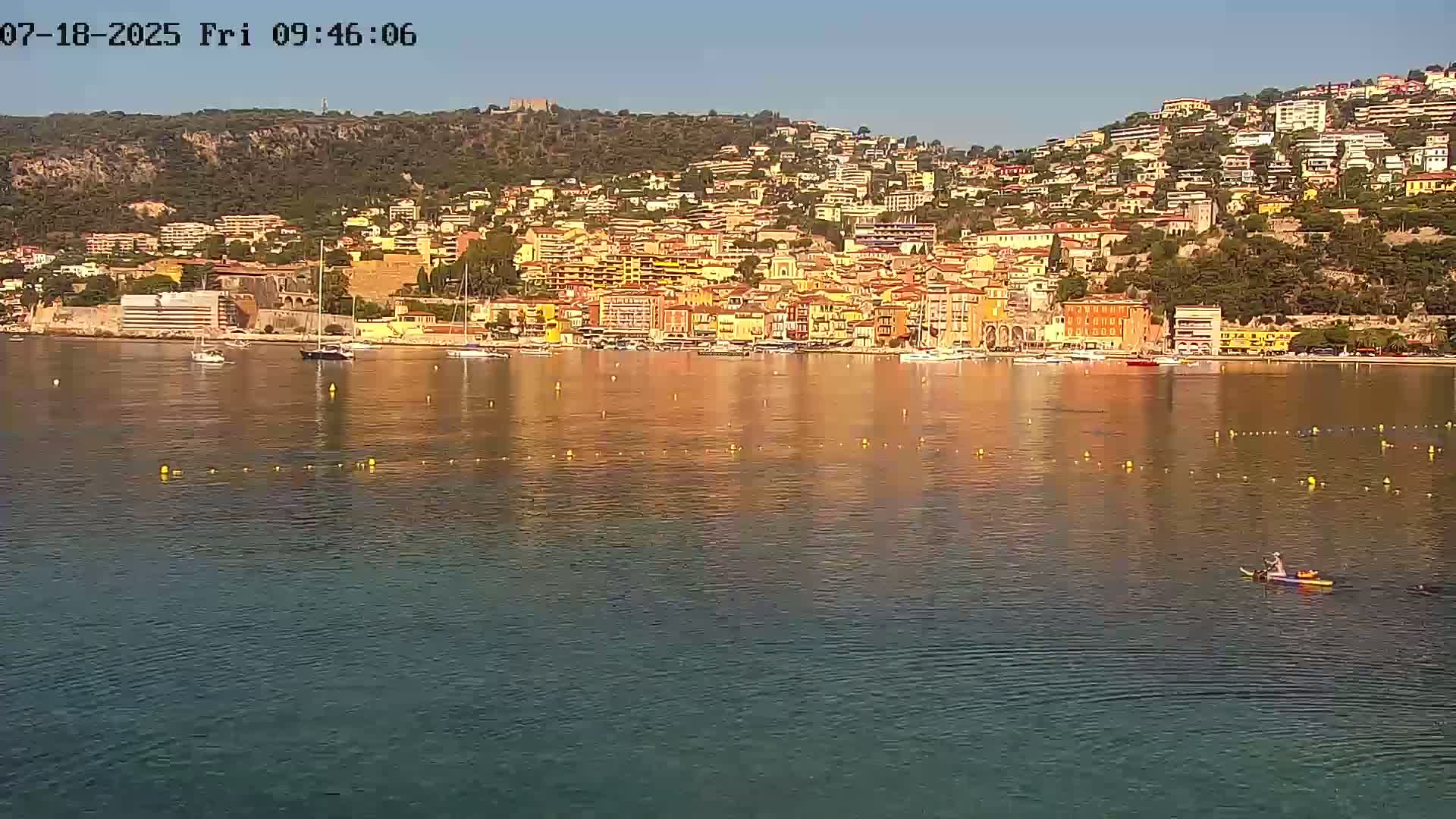 A calm body of water reflects a hillside town of closely packed buildings under a clear sky, with a lone person paddling a small boat in the foreground.