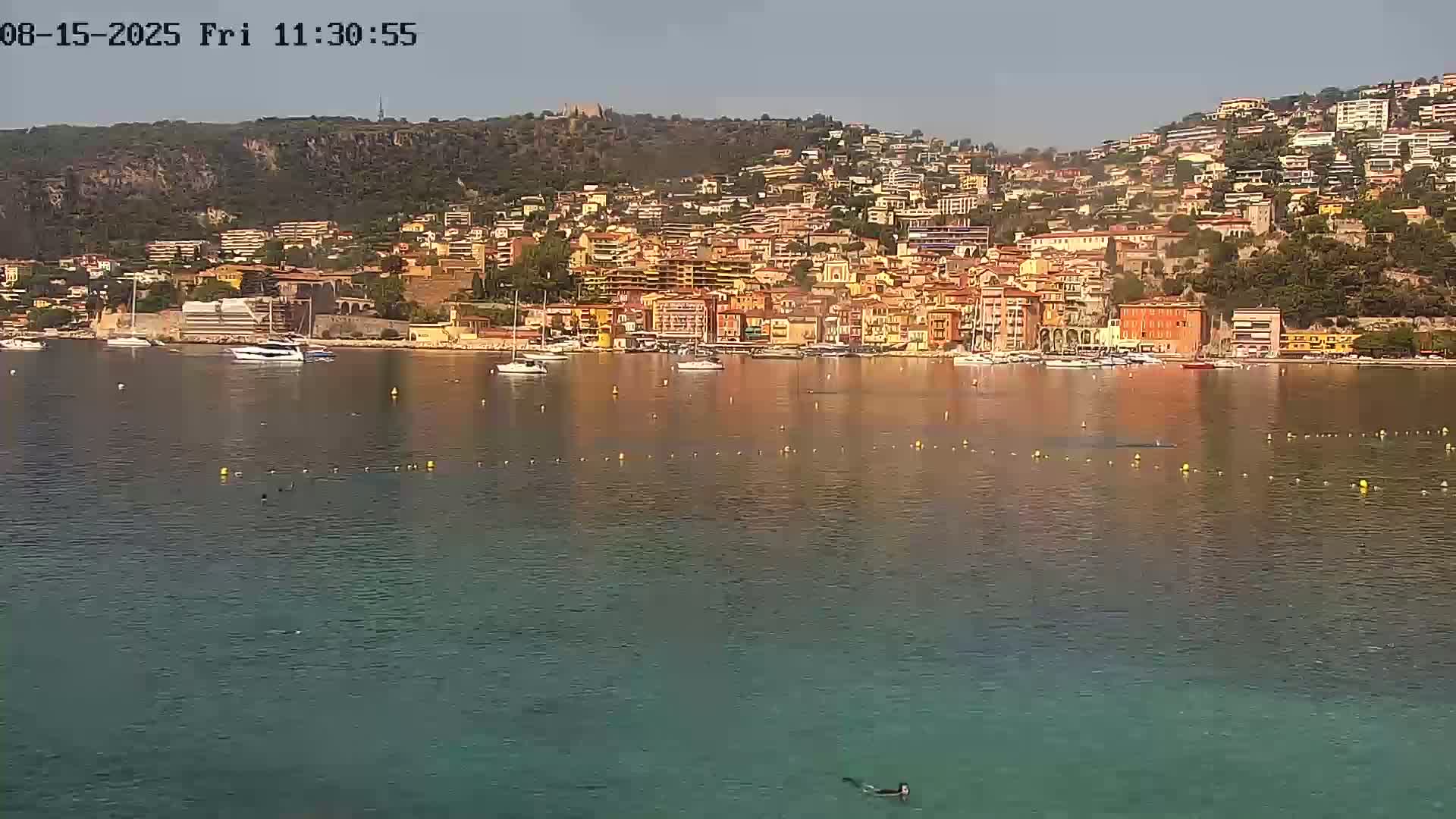 A calm body of water reflects a hillside town of closely packed buildings under a clear sky, with a lone person paddling a small boat in the foreground.