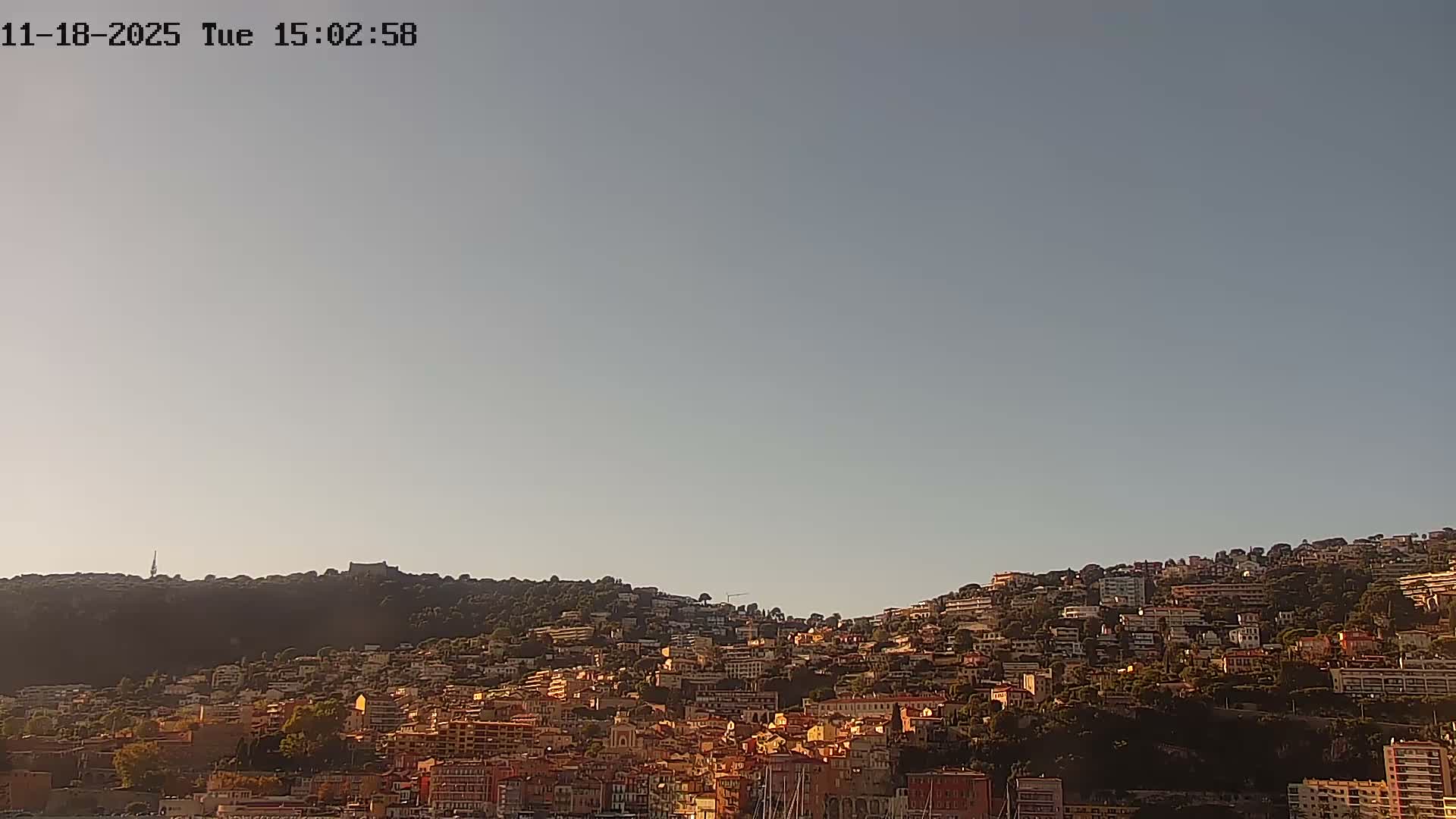 A calm body of water reflects a hillside town of closely packed buildings under a clear sky, with a lone person paddling a small boat in the foreground.