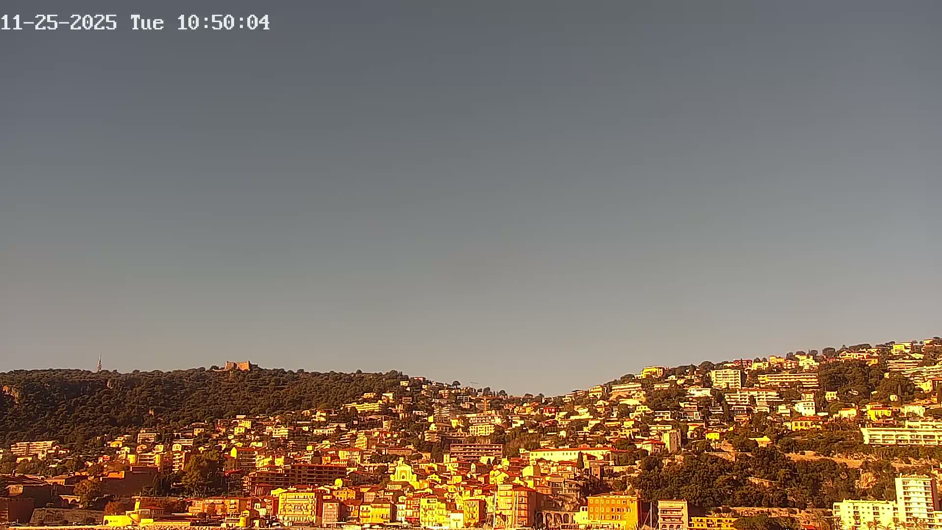 A clear, sunny day illuminates a vibrant, densely built city nestled along a tree-covered hillside beneath a bright blue sky.