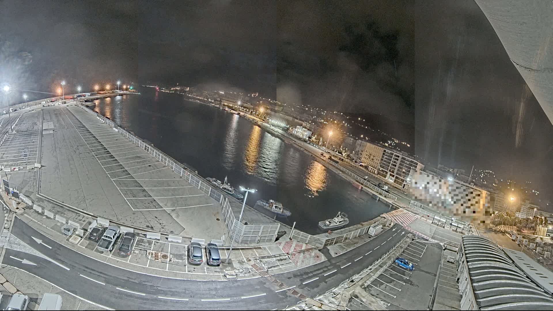 An empty parking lot next to a calm harbor at night, with a few small boats and city lights visible in the distance.
