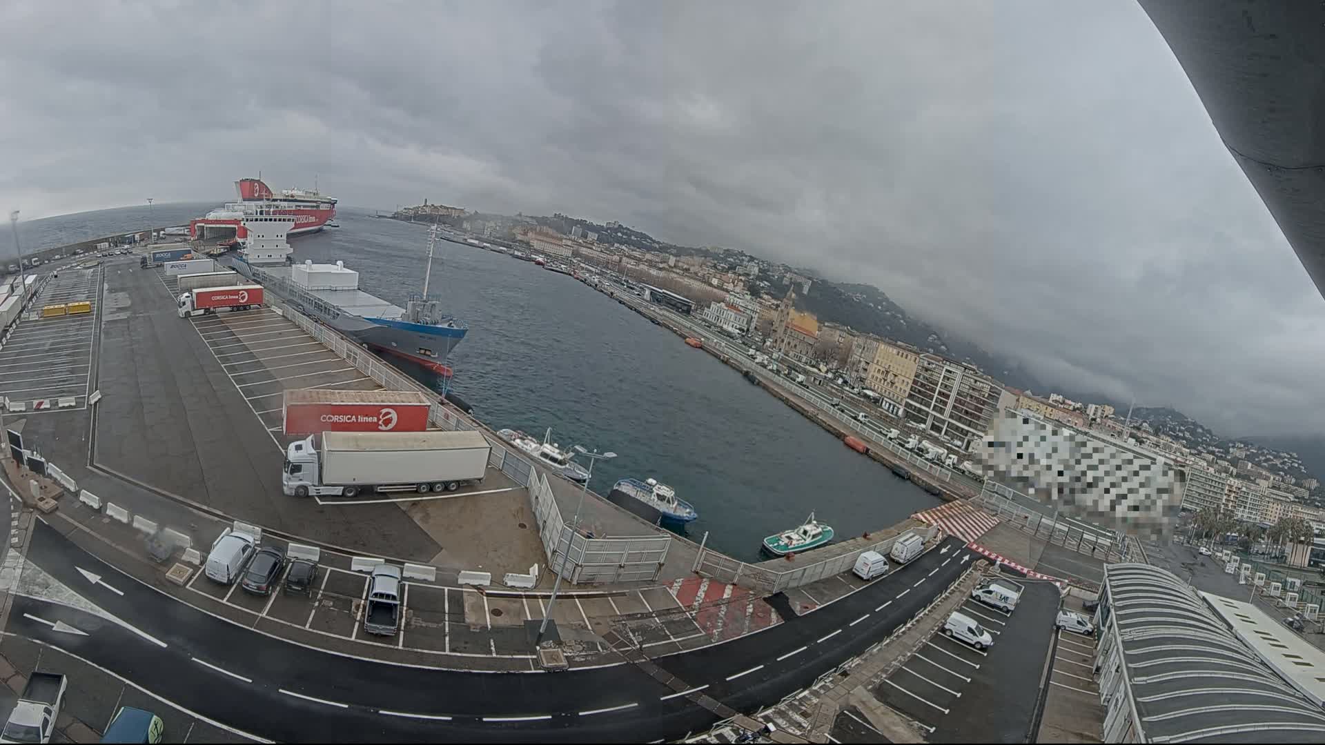 A harbor scene under an overcast sky shows several ships docked, numerous trucks parked, and cars in a parking lot along a waterfront lined with buildings.
