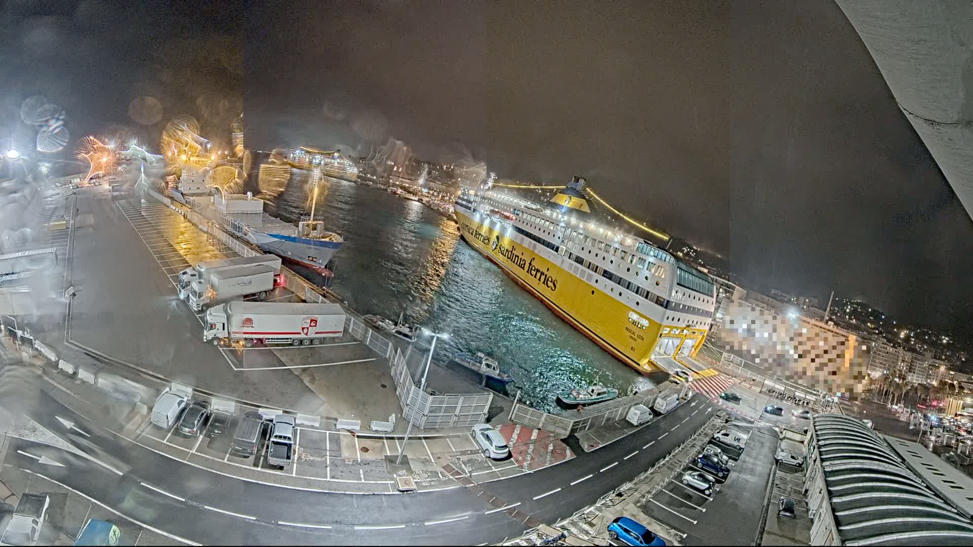 A large yellow ferry is docked at a port at night, with several other ships and trucks in the vicinity, under wet conditions.