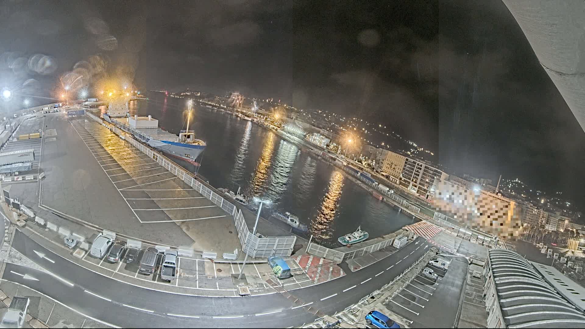 An aerial nighttime view of a harbor with a cargo ship docked, several smaller boats in the water, cars parked in a lot, and city lights illuminating the waterfront.