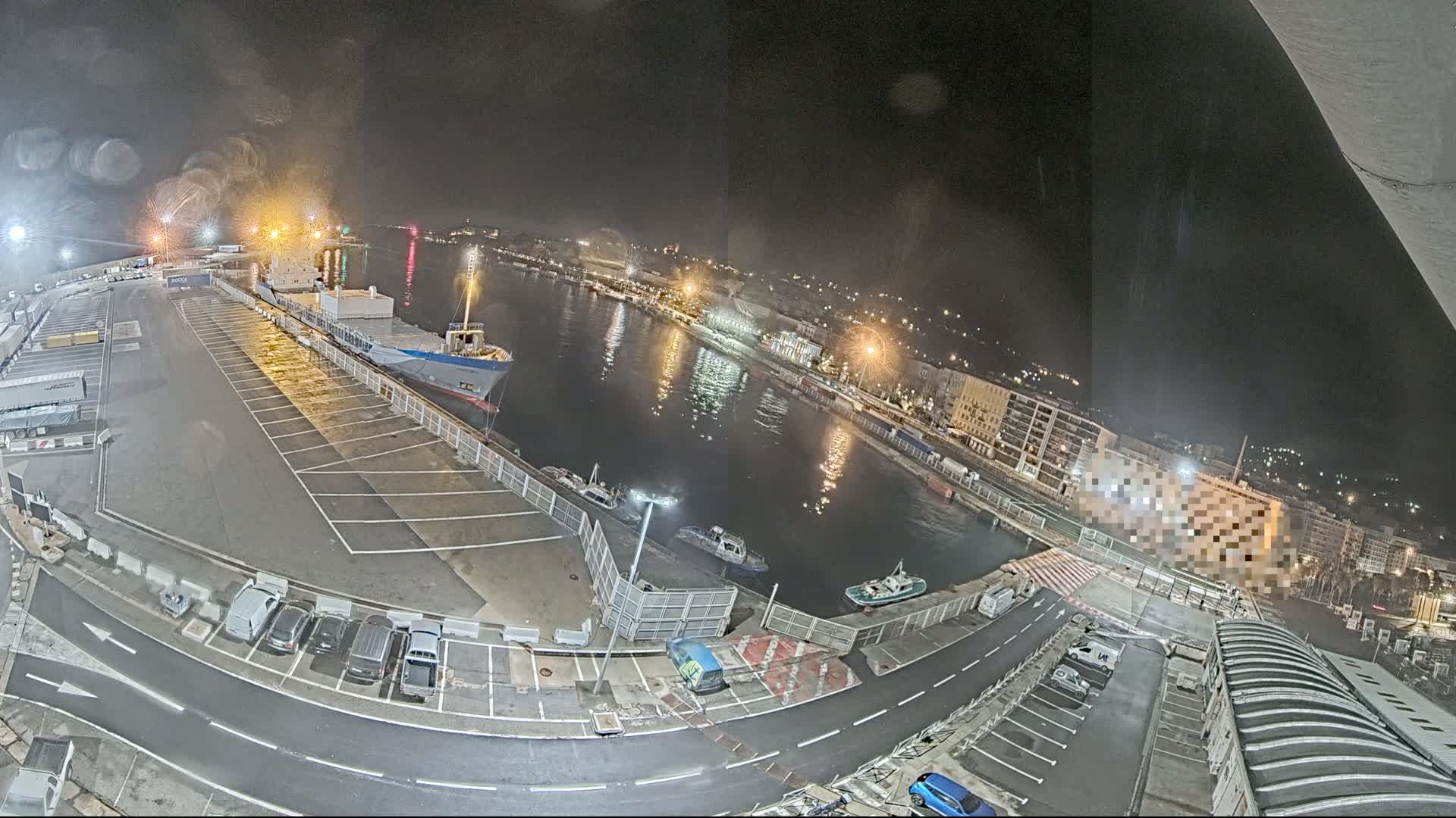 A nighttime aerial view of a harbor, showing a cargo ship docked, several smaller vessels in the water, and a parking lot with cars, all under a dark, possibly slightly rainy sky.