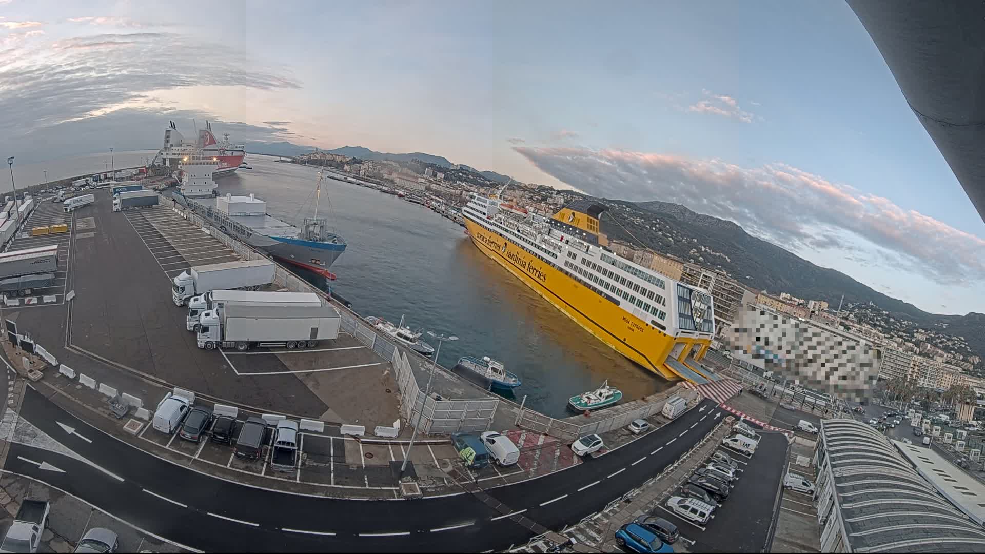 A port with several large ships docked, numerous trucks and cars parked in the surrounding lots, and a city and mountains in the background under a partly cloudy sky.
