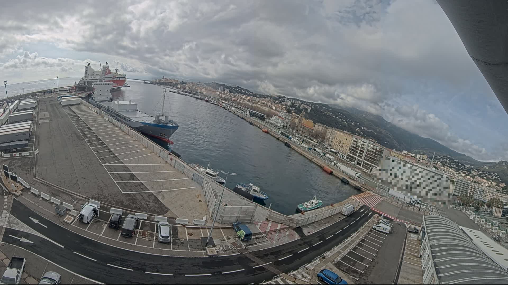 A harbor scene under a cloudy sky shows a large ferry and cargo ship docked, along with several smaller boats and cars parked in a lot.