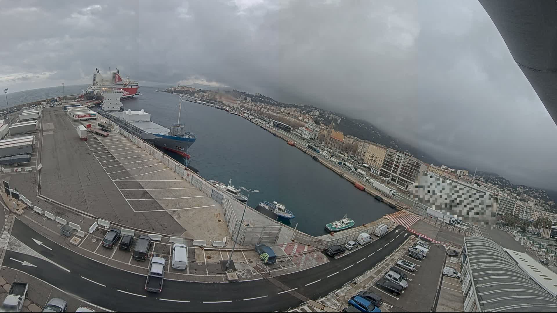 A harbor scene on an overcast day shows several cargo ships and smaller boats docked, with vehicles parked in a nearby lot and a city visible in the background.