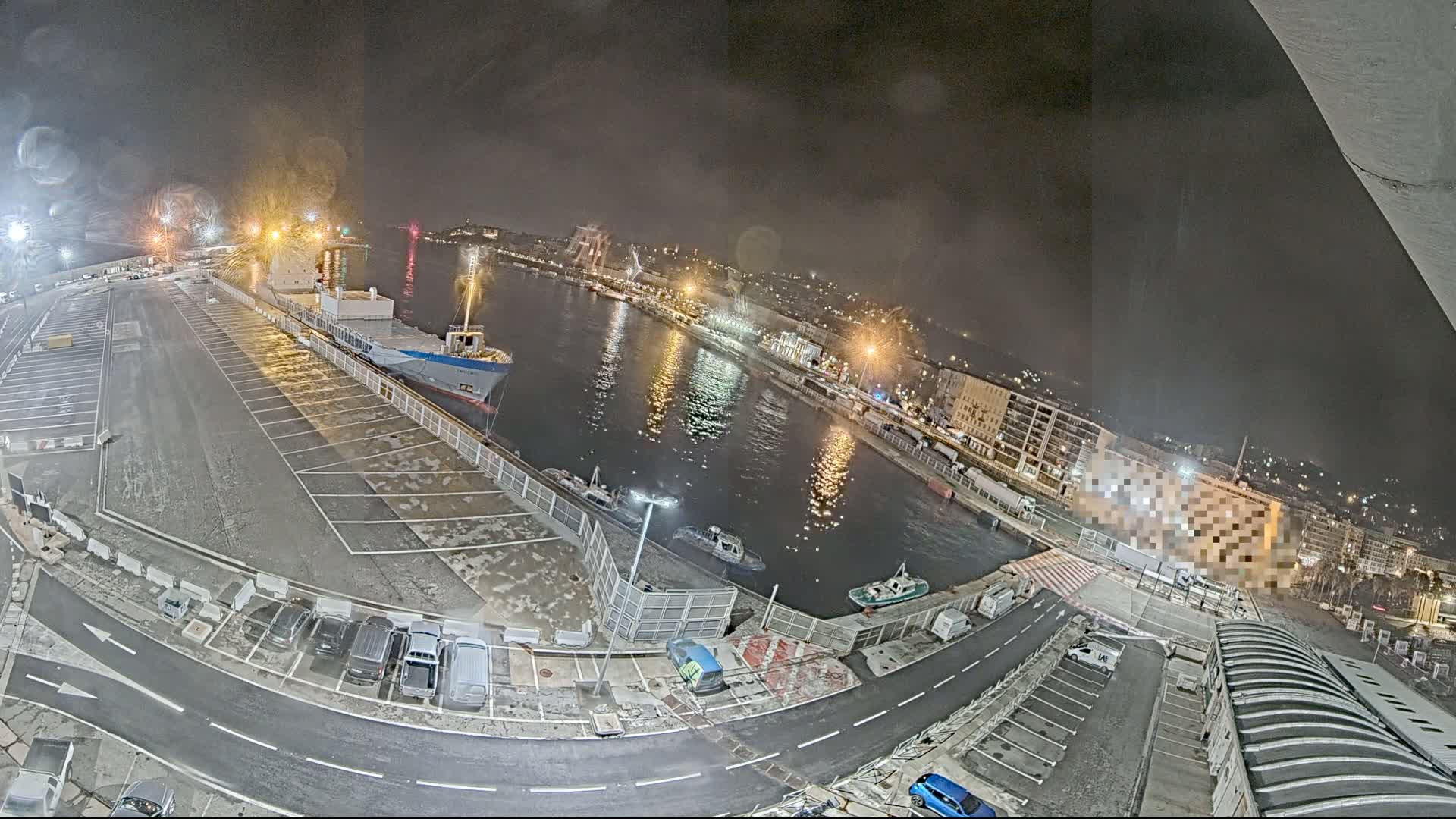 A nighttime aerial view of a harbor with a large cargo ship docked, several smaller boats in the water, and a mostly empty parking lot on the land.