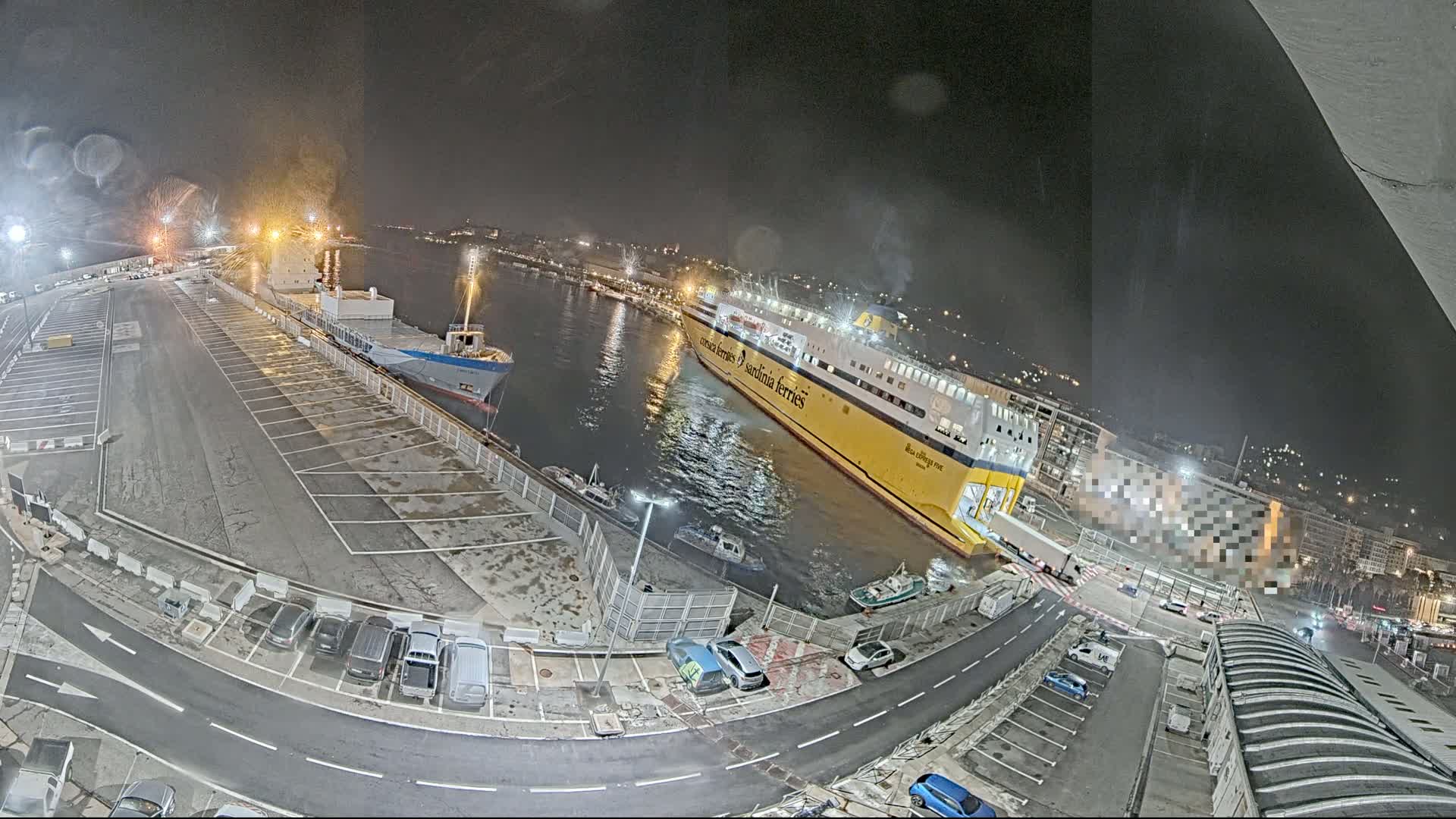 At night, a large yellow ferry is docked next to a smaller cargo ship in a harbor, with cars parked in a nearby lot and other vehicles moving around the port area.