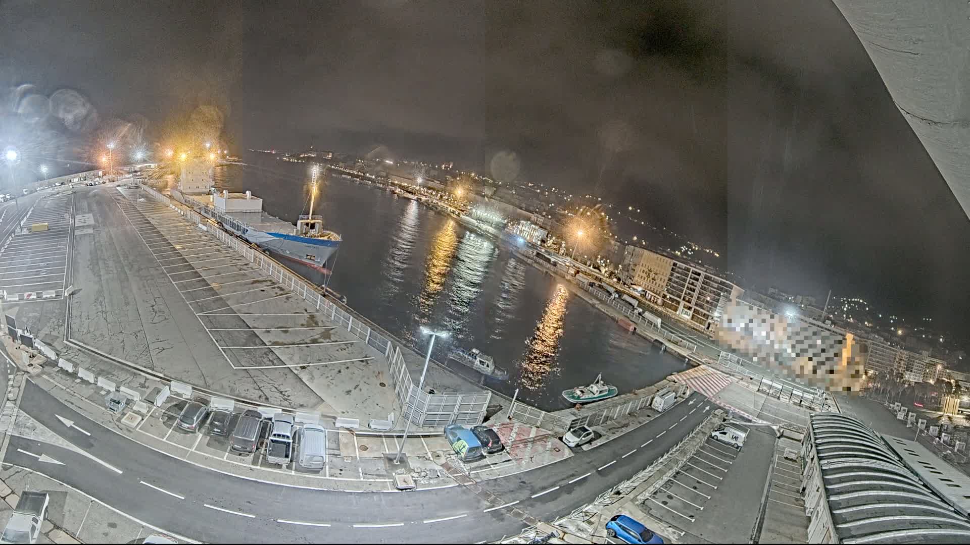 A nighttime aerial view of a harbor with a large cargo ship docked, several smaller boats in the water, and cars parked in lots surrounding the waterway.