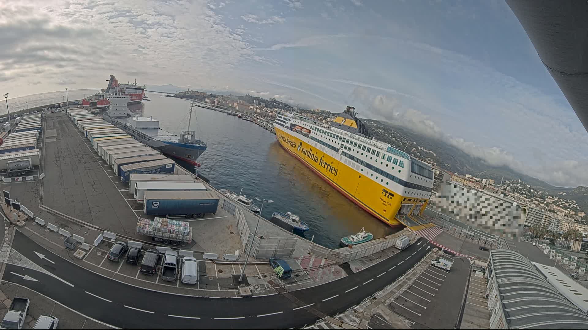 A large yellow ferry is docked at a port alongside several cargo ships and trucks, under a partly cloudy sky.