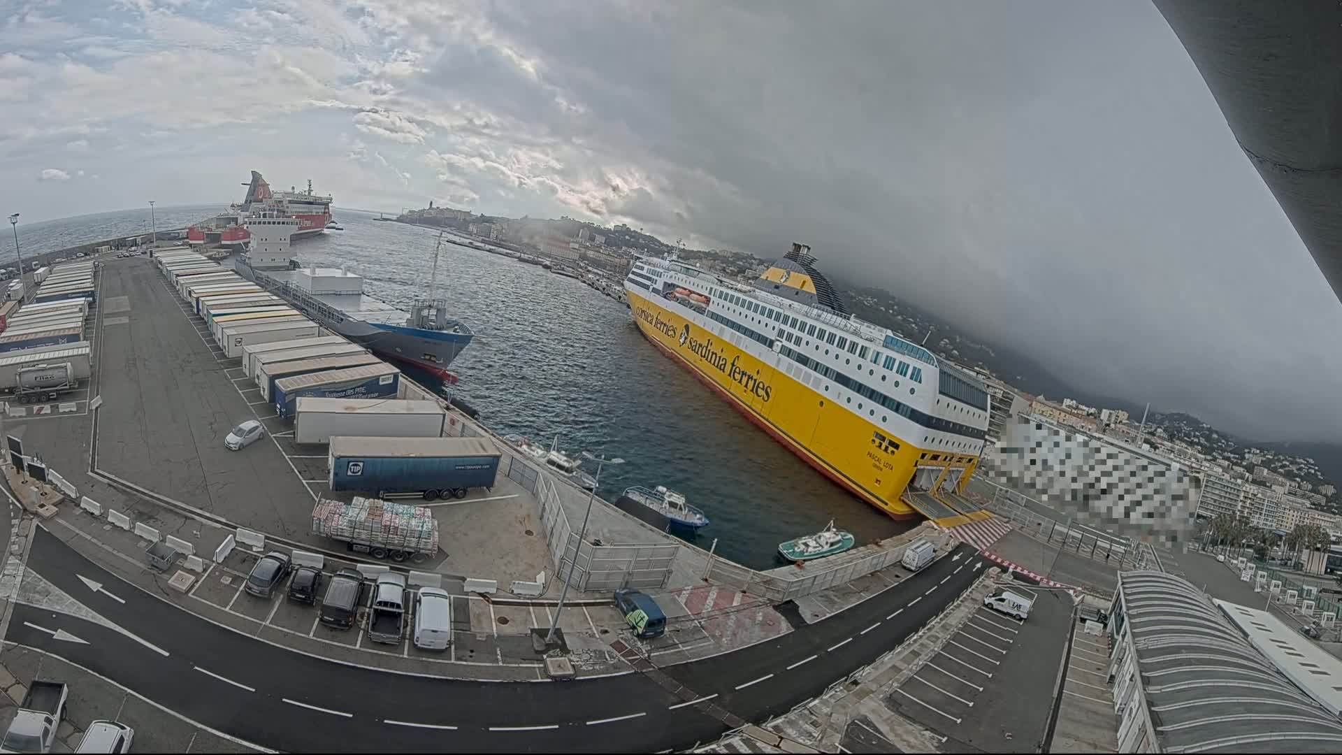 A large yellow ferry is docked at a port under an overcast sky, with other ships and numerous cargo containers visible in the vicinity.