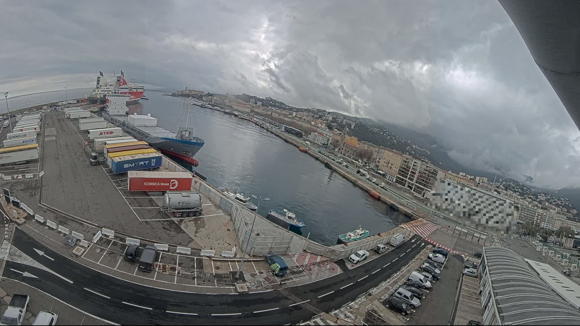 A harbor scene on an overcast day shows cargo ships, trucks, and cars parked around a body of water next to a city.