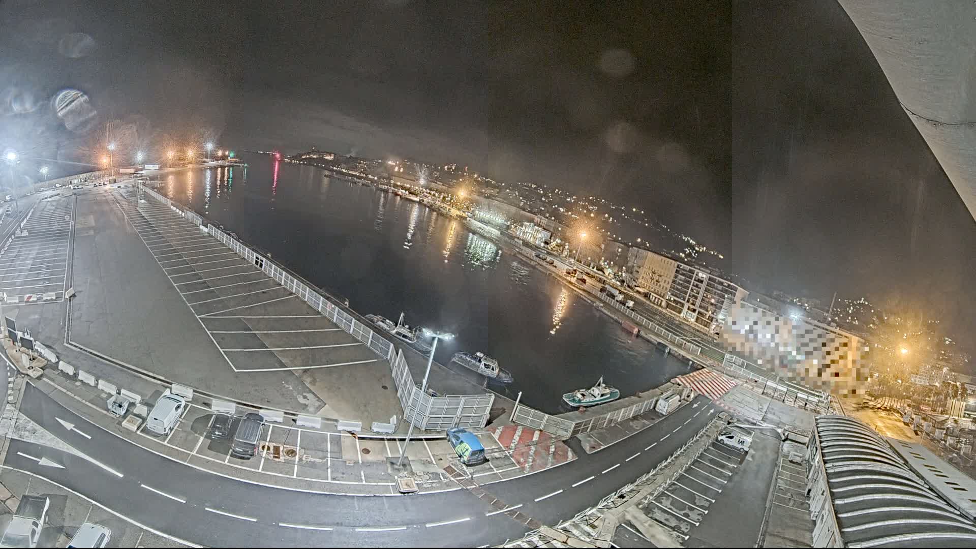 A nighttime aerial view of a harbor with a mostly empty parking lot, several small boats on the water, and city lights in the background under a cloudy sky.
