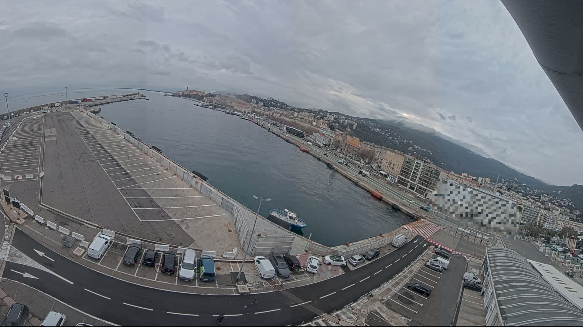 An overcast day reveals a harbor scene with a large empty parking lot, several cars, a small boat in the water, and a city nestled against distant mountains.