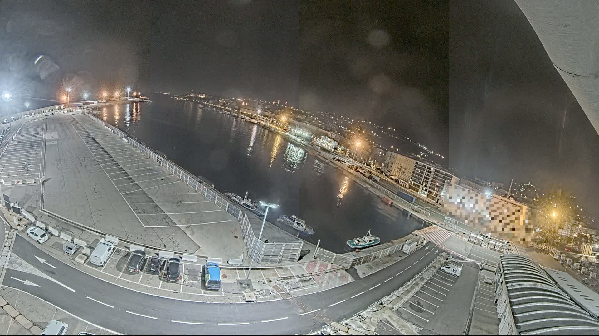 A nighttime aerial view of a harbor, showing a mostly empty parking lot next to a calm body of water with several small boats, and a city illuminated by streetlights in the background.