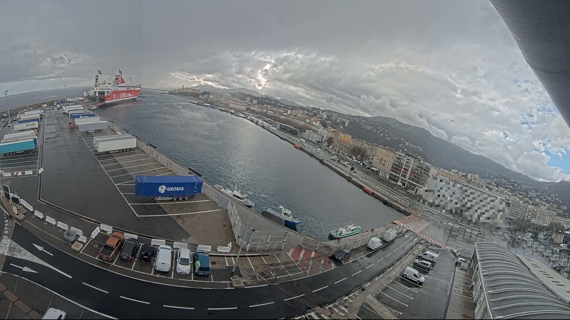 A ferry is docked at a port, next to a parking lot filled with cars and trucks, under an overcast sky.