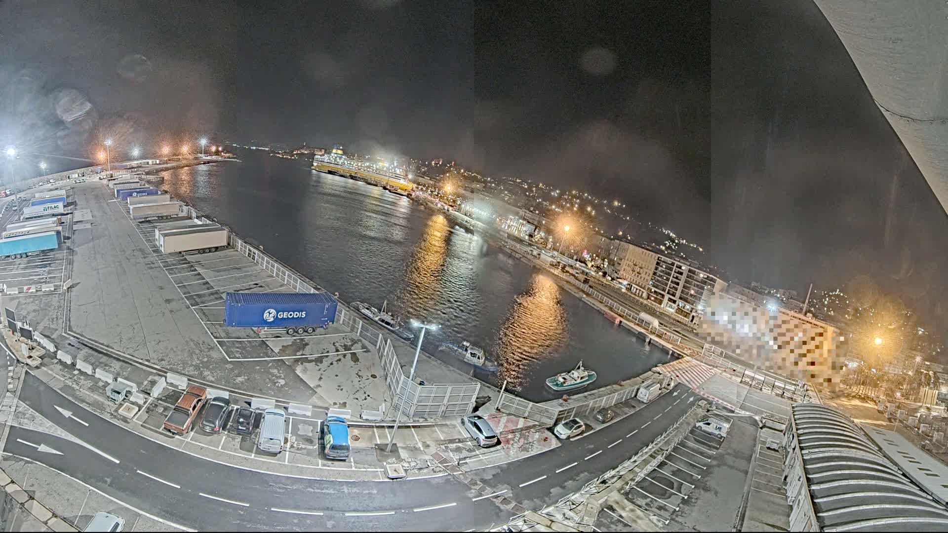 A nighttime aerial view of a harbor with a large ferry docked, numerous parked trucks and cars, and several smaller boats in the water.