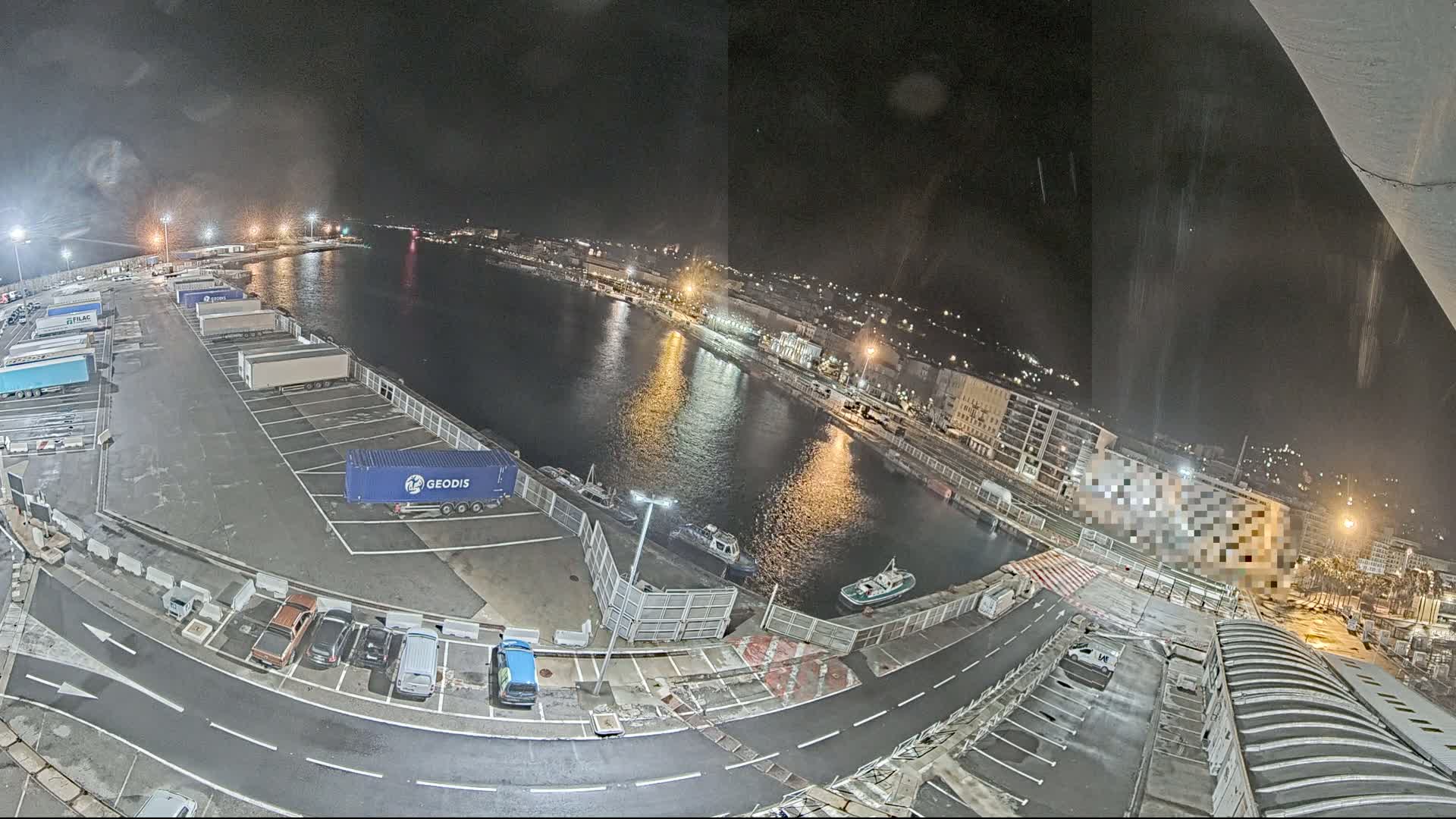 A nighttime aerial view of a harbor shows a waterway reflecting lights from buildings and docked boats, with a parking lot containing several trucks and cars nearby.