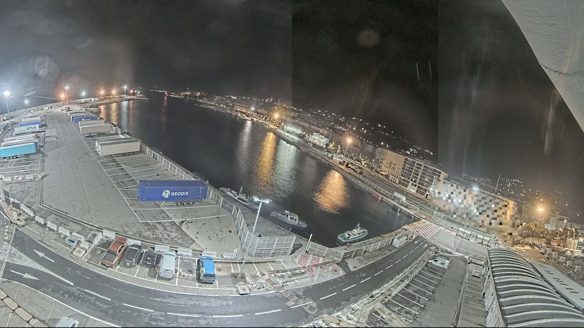 A nighttime aerial view of a harbor with docked boats, parked trucks and cars, and city lights reflected in the calm water.
