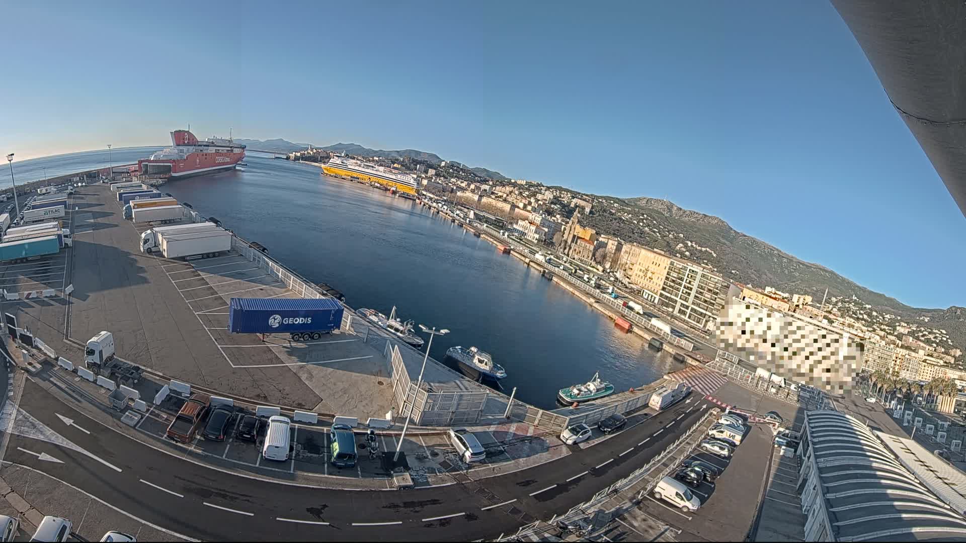 An aerial view of a harbor on a clear, sunny day shows two large ferries docked, numerous trucks and cars parked in lots, and several small boats in the water, all surrounded by buildings and hills.