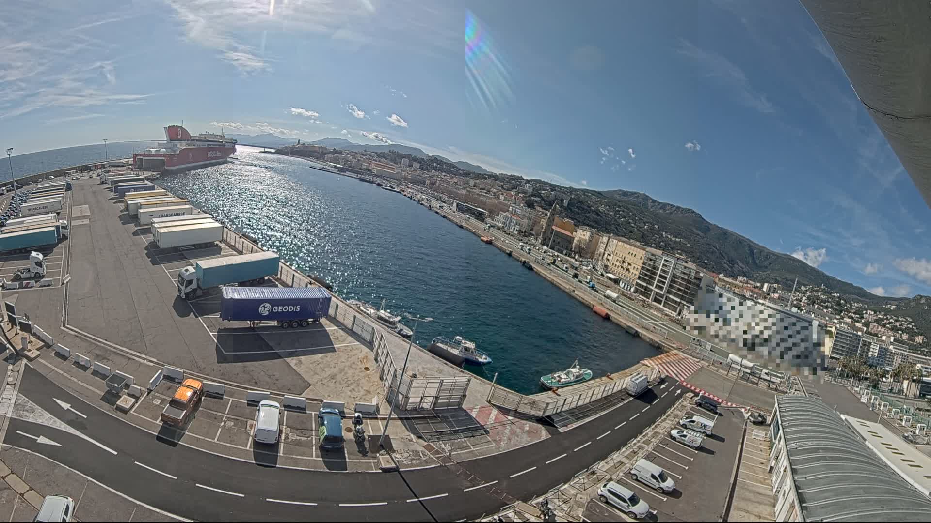 A ferry is docked at a harbor on a sunny day, with numerous trucks and cars parked nearby, and a city and mountains visible in the background.