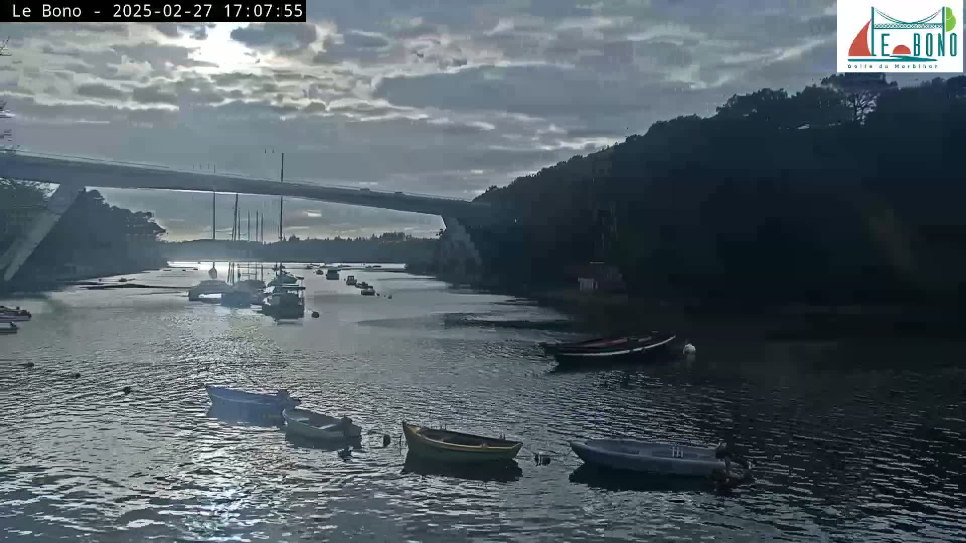 Several small boats are moored in a calm body of water under a cloudy sky, with a long bridge spanning the waterway in the background.