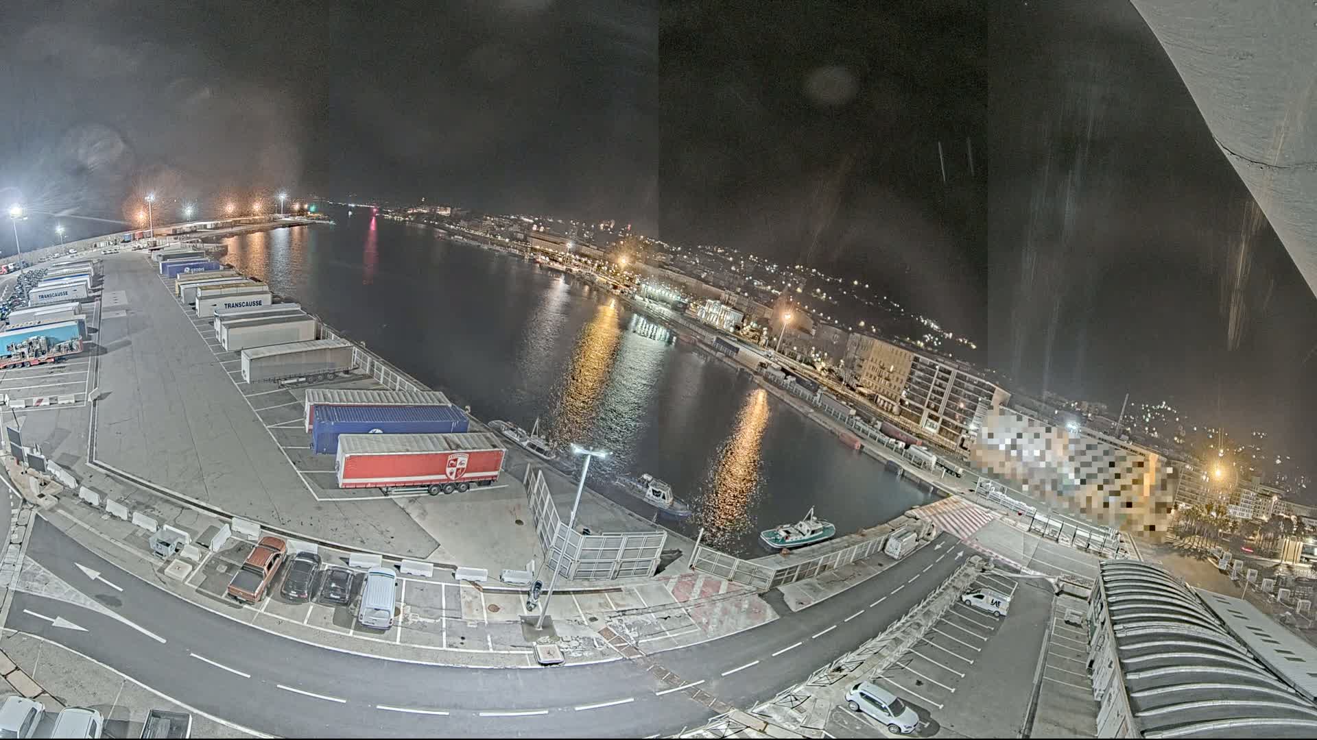 A nighttime aerial view of a harbor at night, showing a parking lot full of semi-trucks and cars near a calm body of water with several small boats.