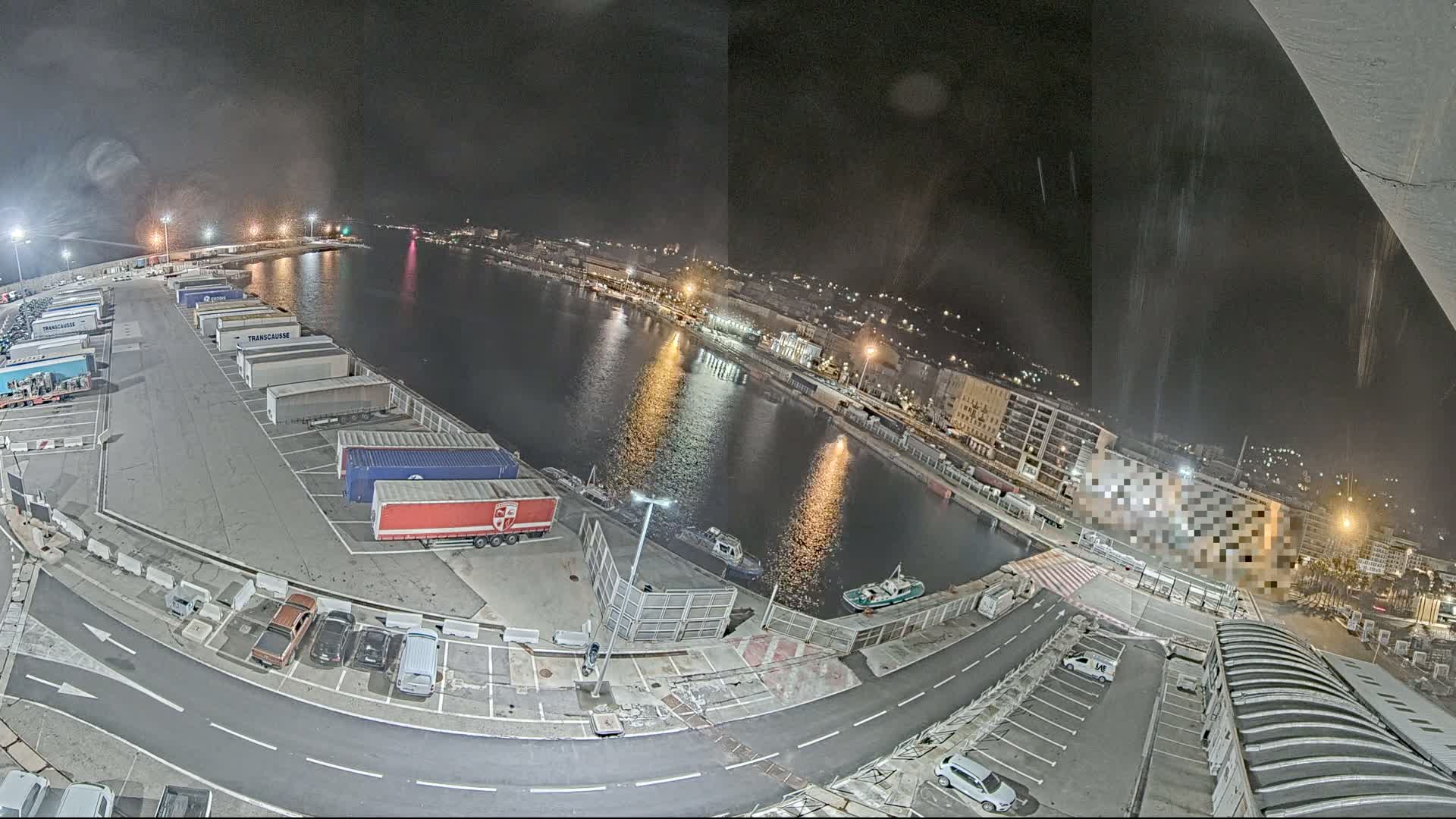 A nighttime aerial view of a harbor with a parking lot full of semi-trailer trucks and cars, a calm body of water reflecting lights from the city, and some small boats.