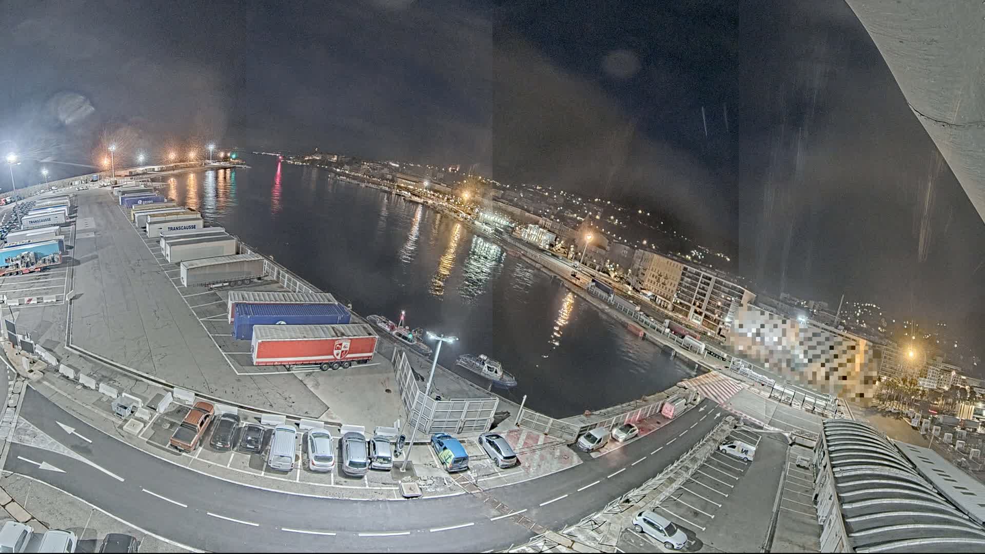 A nighttime aerial view of a harbor, showing a body of water reflecting city lights, with trucks parked in a lot and cars in another lot nearby.