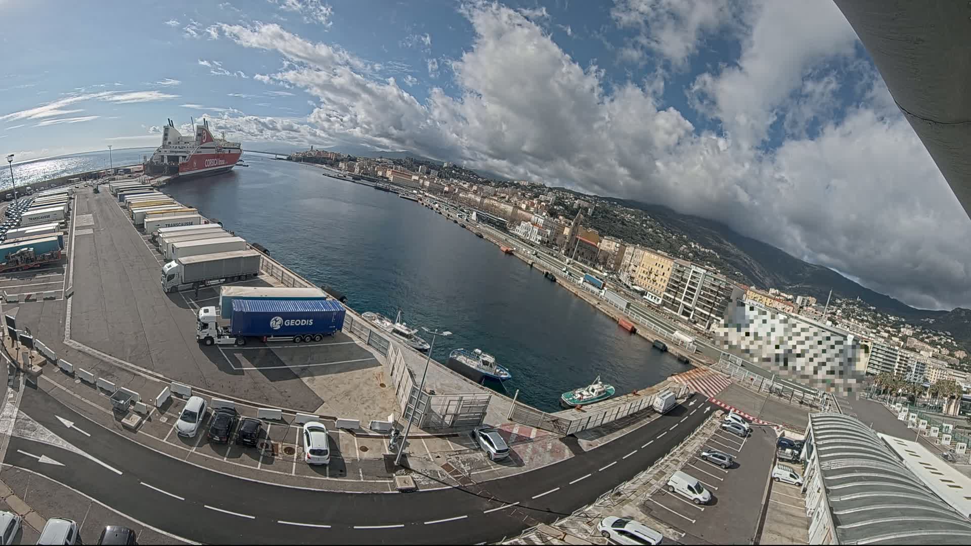 A large ferry is docked at a harbor in a city on a partly cloudy day, with numerous trucks and cars parked nearby.