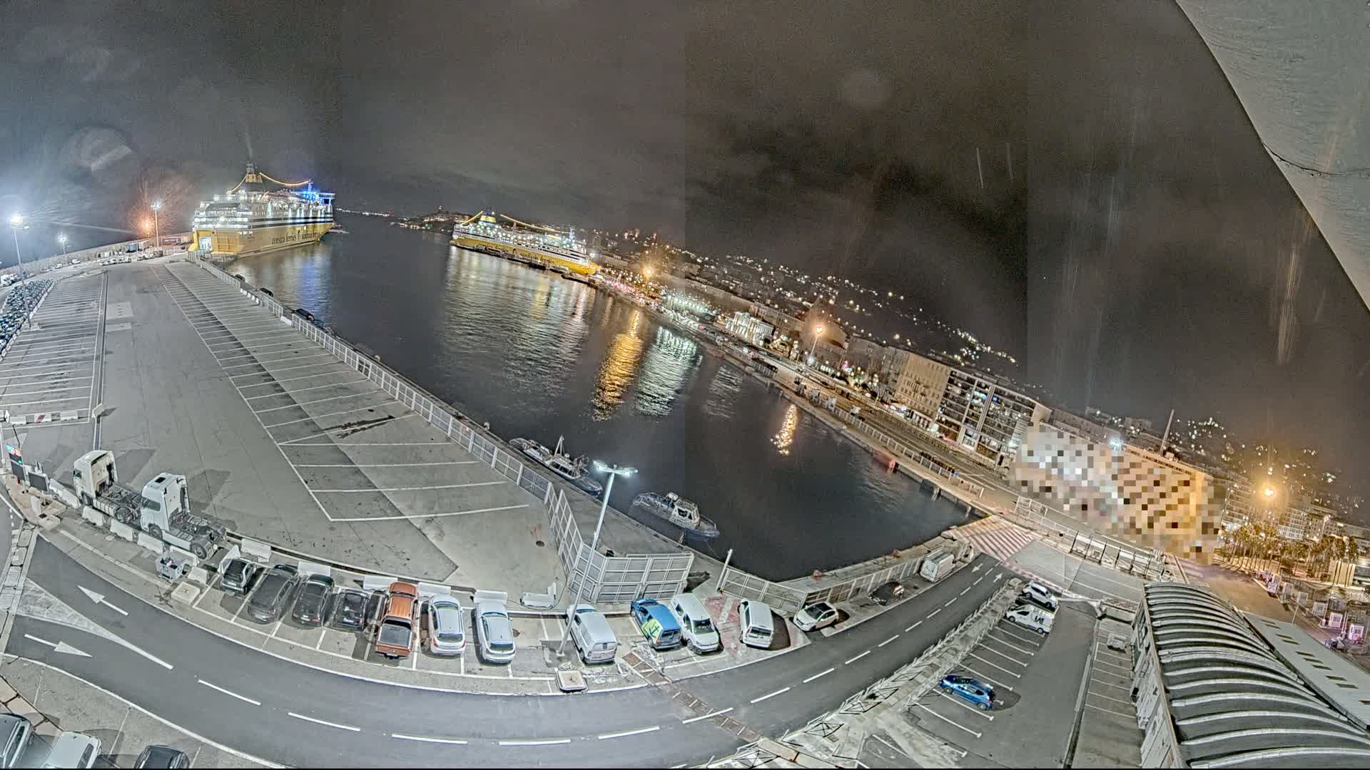 At night, a harbor is shown with two large ferries docked, several cars parked in lots surrounding the water, and a city in the background under a dark, possibly rainy sky.