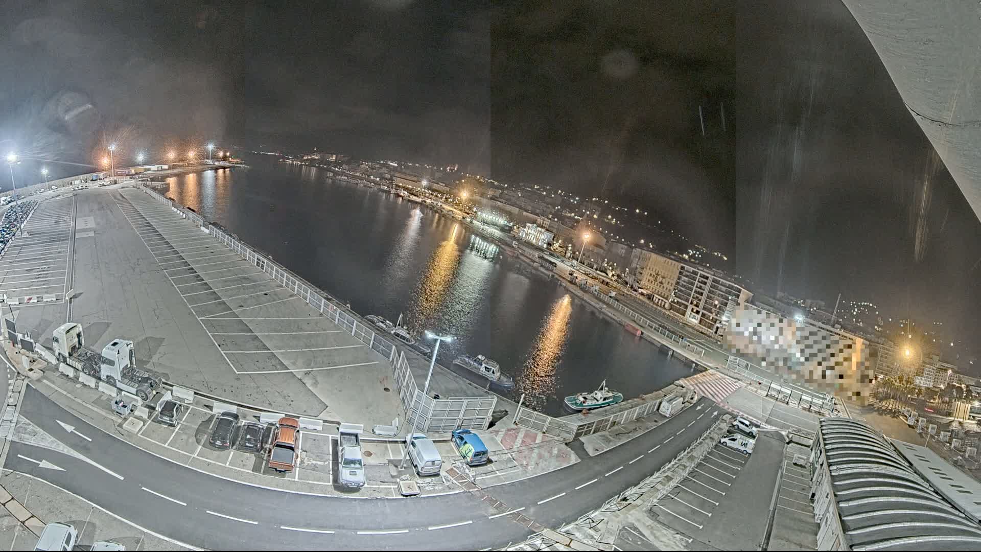A nighttime aerial view of a harbor with a large empty parking lot, several small boats on the water, and buildings illuminated along the shoreline.