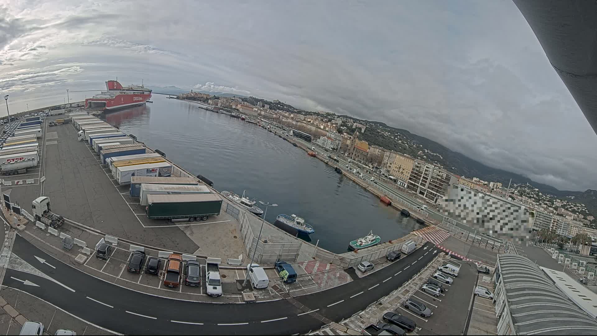 A large red ferry is docked at a harbor, with numerous trucks parked in a lot next to it, under an overcast sky.