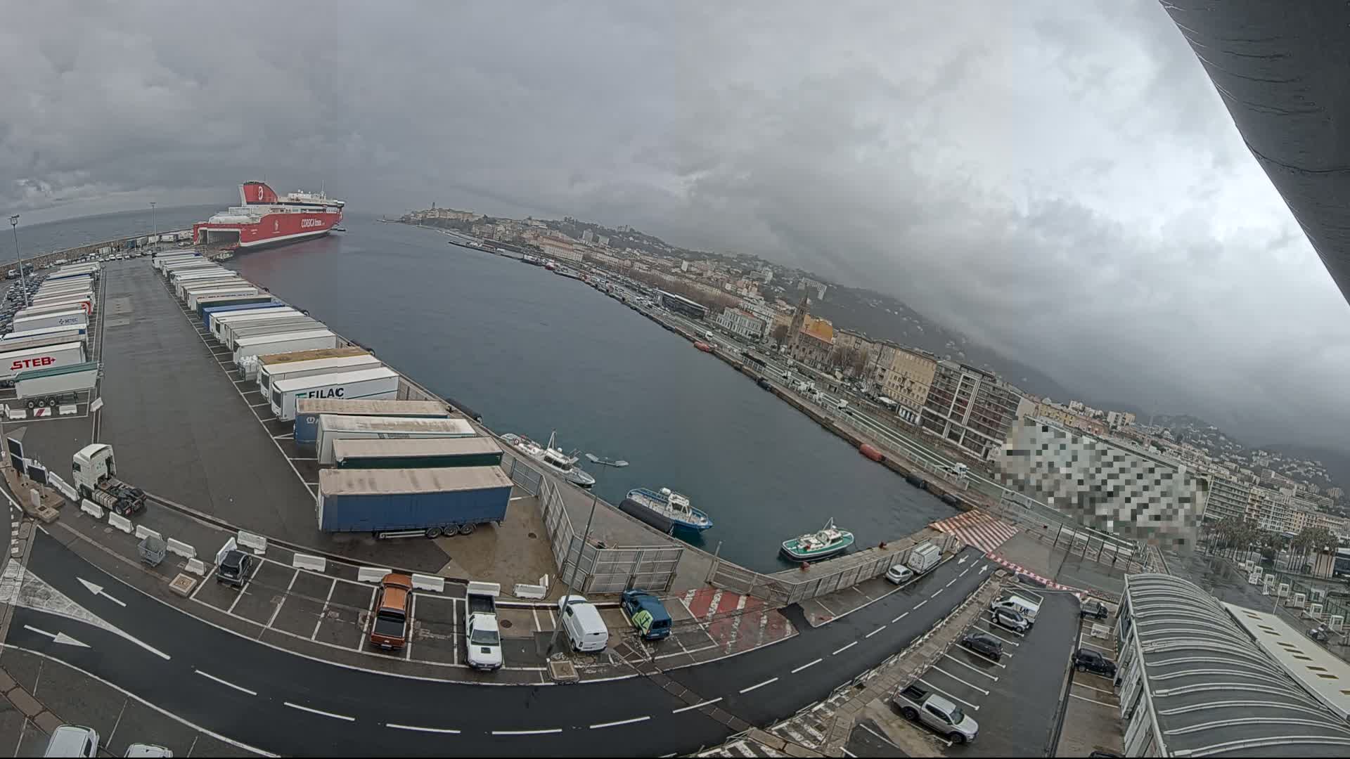 A large red ferry is docked at a port, numerous trucks and cars are parked in the lot, and smaller boats are on the water under an overcast sky.