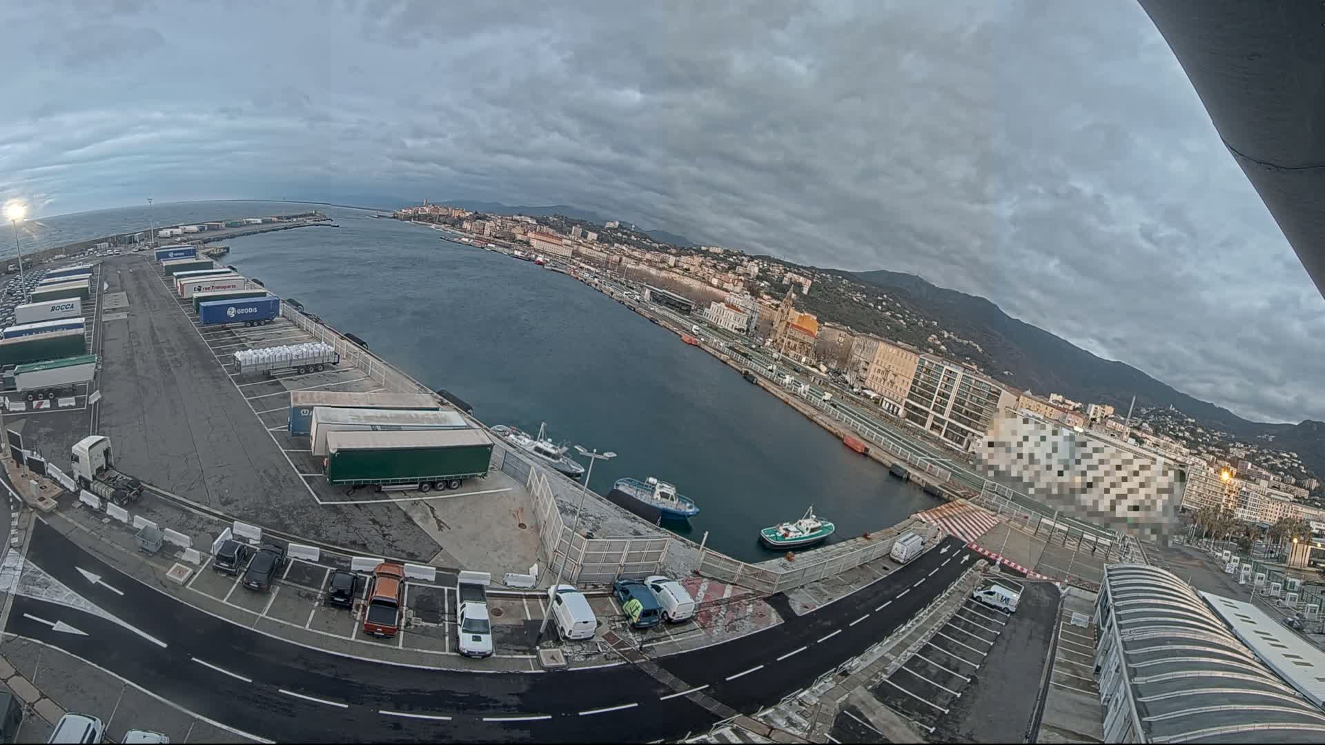 An overcast day at a harbor shows a large parking lot filled with semi-trucks, several smaller vehicles, and a calm body of water with several small boats.