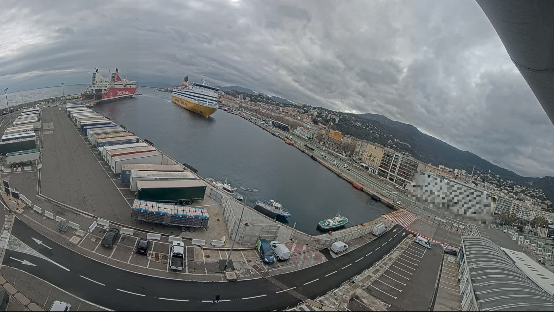 An overcast day at a harbor shows two large ferries docked, numerous trucks parked in a lot, several smaller boats in the water, and a city with mountains in the background.