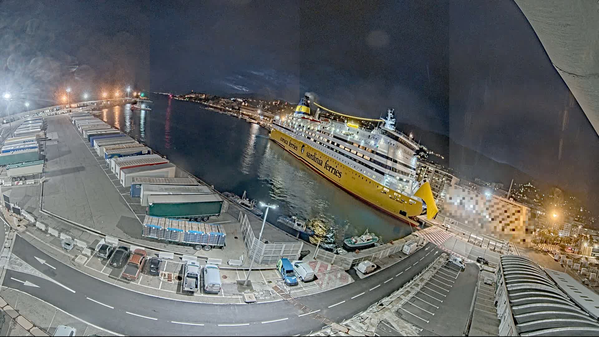 A large yellow ferry is docked at a port at night, surrounded by trucks, cars, and other vehicles parked in a lot.