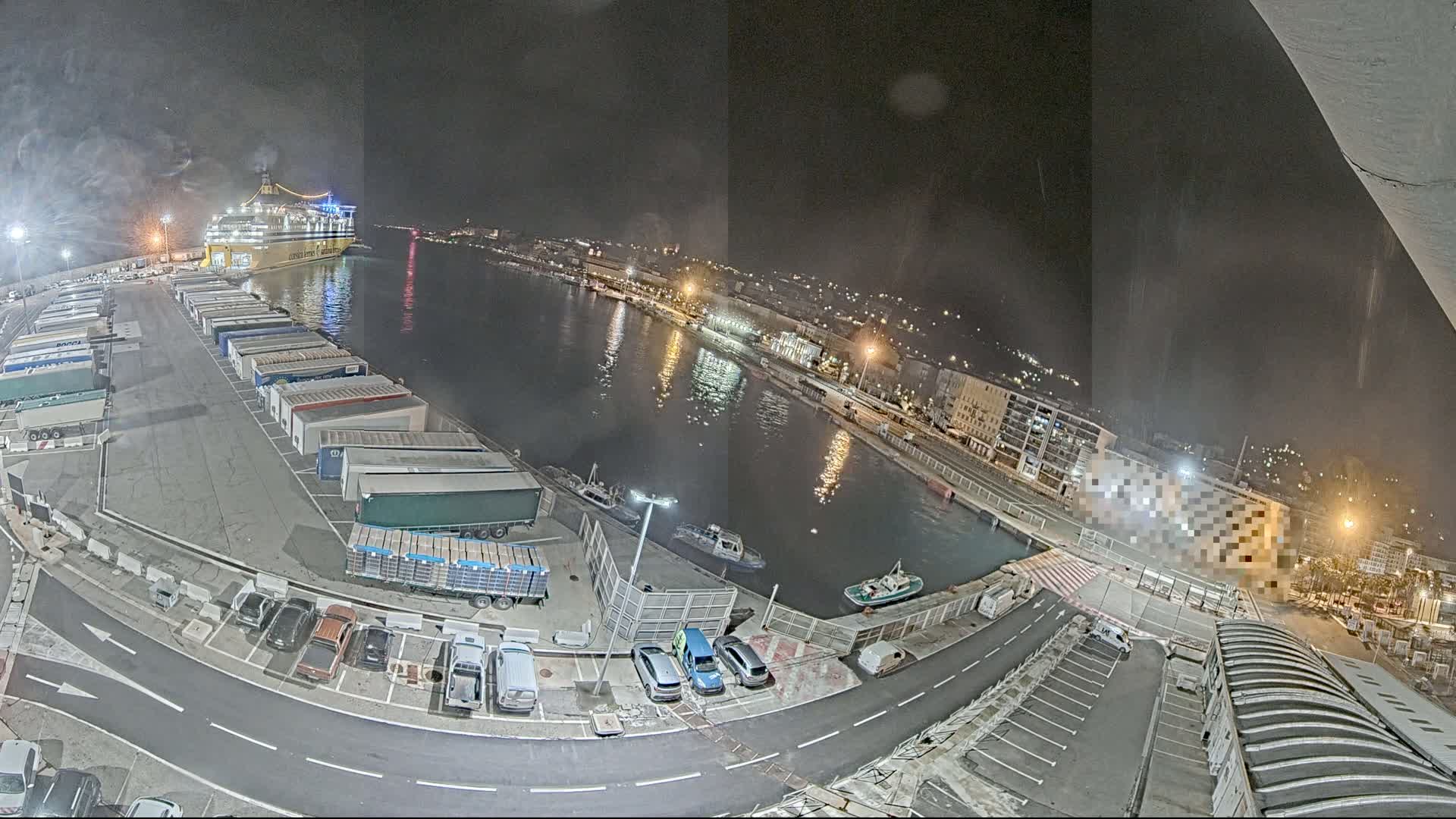A nighttime aerial view of a harbor shows a large ferry docked near numerous trailers and a few cars parked in a lot, with small boats in the water.